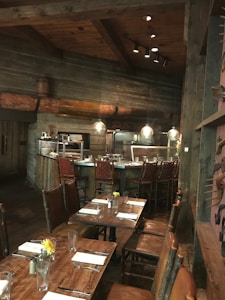 A rustic dining area with wooden tables and chairs, set up for service. The tables are adorned with glassware, silverware, folded napkins, and small floral arrangements in jars. The setting features wooden walls and ceiling beams, with low-hanging pendant lights providing warm illumination. In the background, there is a bar area with high-backed woven bar stools.
