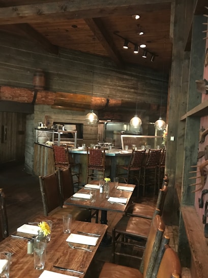 A rustic dining area with wooden tables and chairs, set up for service. The tables are adorned with glassware, silverware, folded napkins, and small floral arrangements in jars. The setting features wooden walls and ceiling beams, with low-hanging pendant lights providing warm illumination. In the background, there is a bar area with high-backed woven bar stools.