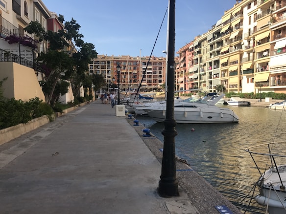 A scenic canal with several yachts docked along the waterway. The promenade is lined with trees and colorful buildings, creating an inviting atmosphere. People are walking along the path, enjoying the sunny day.