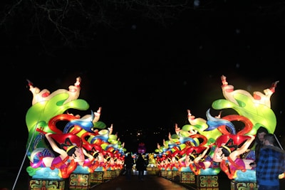 A series of illuminated, colorful lantern sculptures depicting stylized figures and elaborate designs, set against a dark background. The lanterns feature vibrant greens, reds, blues, and yellows, with swirling elements and multiple figures arranged symmetrically on either side of a pathway. Two people are visible in the foreground, admiring the display.