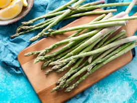Fresh spears of asparagus are laid out on a wooden cutting board with a blue cloth underneath. A dish containing lemon wedges is visible in the top left corner, set against a turquoise textured surface.