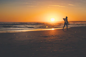 A happy surfer carrying a stand-up paddleboard along the shore at sunset.