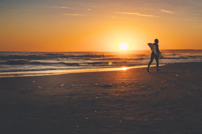 A happy surfer carrying a stand-up paddleboard along the shore at sunset.
