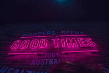Neon pink letters spell out 'GOOD TIMES' against a dark background with faint, partially visible words reading 'BURGERS, BEERS' and '100% AUSTRALIAN'. The ambiance gives a retro, energetic vibe often found in restaurants or bars.