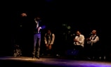 A group of performers is on stage in a dimly lit setting. One person, standing in the foreground, appears to be dancing or gesturing expressively. Three others are seated in the background, dressed in formal attire, with microphones in front of them, clapping along to the rhythm.