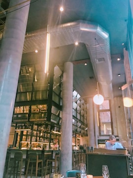 A modern industrial-style bar area with high ceilings featuring large pillars and exposed ventilation ducts. The bar is well-stocked with bottles and glasses, and there are several high chairs lined along it. Warm lighting from pendant lights softens the atmosphere. A person is present, seated at a table, seemingly reading or working.