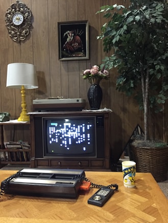 A vintage living room setup featuring a wood-paneled wall with decorative elements such as a clock and a painting. A CRT television displays a retro video game, likely from an early console. On the table in front of the TV are an old gaming console or device with a connected joystick and a cup with a cartoon character design. A lamp with a yellow base, a vinyl record player, a vase with flowers, and a large indoor plant are also visible in the room.
