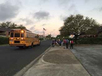 A yellow school bus is stopped on a suburban street, with a group of students walking down the sidewalk. The sky is partly cloudy with a hint of sunset, and an American flag is visible in the background. A large tree and residential houses line the street.