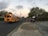 A yellow school bus is stopped on a suburban street, with a group of students walking down the sidewalk. The sky is partly cloudy with a hint of sunset, and an American flag is visible in the background. A large tree and residential houses line the street.