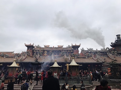 A traditional temple with intricate architectural details and ornate rooftops, bustling with people, likely visitors or worshippers, engaged in various activities. Smoke is emanating from the temple area, suggestive of incense burning or rituals taking place.