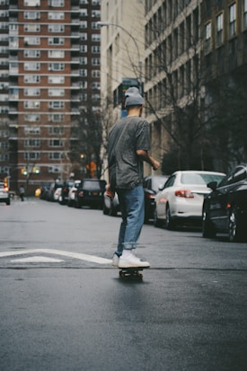 A person skateboards down a wet city street, surrounded by parked cars and high-rise buildings. They wear a beanie, striped shirt, jeans, and white sneakers. The urban setting includes overcast skies and bare trees, typical of a cold season.