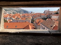 A picturesque view of a historic cityscape featuring red-tiled roofs and stone buildings. The image is framed by a stone window opening, providing a sense of looking out over the city. In the distance, there are hills and clear skies, adding to the charming and tranquil atmosphere.