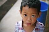 A young child with wide eyes and a curious expression is wearing a plaid shirt. The background is slightly blurred, with a large blue object, possibly a container, visible to the right.