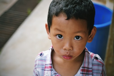 A young child with wide eyes and a curious expression is wearing a plaid shirt. The background is slightly blurred, with a large blue object, possibly a container, visible to the right.
