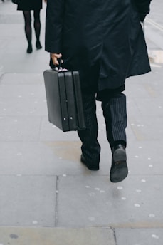 An individual dressed in formal attire, wearing a long black coat and pinstriped trousers, is walking on a gray pavement holding a black briefcase in their right hand. Another person is visible ahead, also dressed in dark clothing.