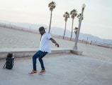 A person is dancing energetically on a beachside walkway. The individual is wearing a white shirt, blue jeans, red sneakers, and a cap. Nearby is a speaker, suggesting music is playing. In the background, there are palm trees and a sandy beach, with mountains visible in the distance.