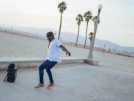 A person is dancing energetically on a beachside walkway. The individual is wearing a white shirt, blue jeans, red sneakers, and a cap. Nearby is a speaker, suggesting music is playing. In the background, there are palm trees and a sandy beach, with mountains visible in the distance.