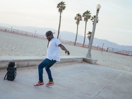 A person is dancing energetically on a beachside walkway. The individual is wearing a white shirt, blue jeans, red sneakers, and a cap. Nearby is a speaker, suggesting music is playing. In the background, there are palm trees and a sandy beach, with mountains visible in the distance.