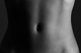A close-up black and white photograph of a human torso focused on the abdominal area. The image emphasizes the natural shadows and contours of the skin, highlighting the texture and shape around the navel.