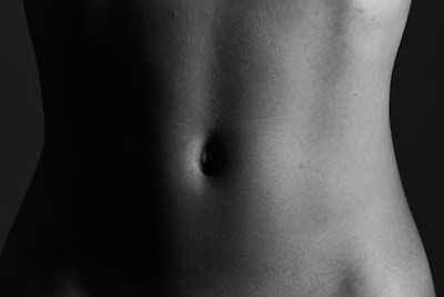 A close-up black and white photograph of a human torso focused on the abdominal area. The image emphasizes the natural shadows and contours of the skin, highlighting the texture and shape around the navel.