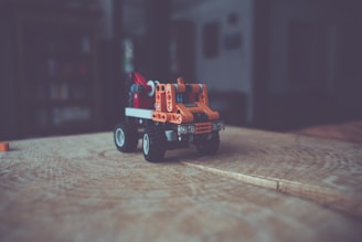 A small toy vehicle made from interlocking plastic bricks, primarily orange, sits on a wooden surface. The background is blurred, creating a sense of depth, and suggesting an indoor setting.