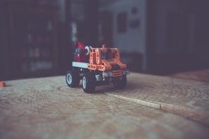 A small toy vehicle made from interlocking plastic bricks, primarily orange, sits on a wooden surface. The background is blurred, creating a sense of depth, and suggesting an indoor setting.