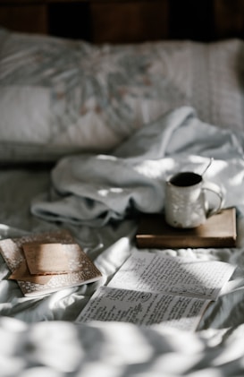 A cozy scene featuring a bed with a white, patterned pillow and soft bedding. Sunlight filters through, creating dappled shadows across handwritten notes and an open journal. A mug with a spoon rests on top of a closed book, suggesting a relaxed moment of reflection or creativity.