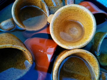 Several brown ceramic cups are partially submerged in clear water, revealing glossy surfaces with reflections. One cup is prominently tilted, while others are arranged at different angles. The colors range from earthy browns to hints of dark reds and greens.