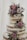 A rustic, three-tiered naked cake adorned with delicate purple and pink roses accompanied by small sprigs of white and purple flowers. Fresh green foliage adds contrast to the pastel blooms, and the cake is lightly dusted with powdered sugar. The soft tones of the flowers and minimalist design create an elegant and romantic feel.