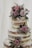 A rustic, three-tiered naked cake adorned with delicate purple and pink roses accompanied by small sprigs of white and purple flowers. Fresh green foliage adds contrast to the pastel blooms, and the cake is lightly dusted with powdered sugar. The soft tones of the flowers and minimalist design create an elegant and romantic feel.