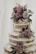 A rustic, three-tiered naked cake adorned with delicate purple and pink roses accompanied by small sprigs of white and purple flowers. Fresh green foliage adds contrast to the pastel blooms, and the cake is lightly dusted with powdered sugar. The soft tones of the flowers and minimalist design create an elegant and romantic feel.