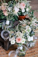 A collection of floral arrangements featuring pastel-colored roses, white flowers, and various greenery. The flowers are arranged in glass jars and a small terracotta pot, placed on a wooden pallet. The setting appears to be indoors with a woven rug underneath.