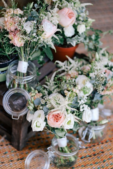 A collection of floral arrangements featuring pastel-colored roses, white flowers, and various greenery. The flowers are arranged in glass jars and a small terracotta pot, placed on a wooden pallet. The setting appears to be indoors with a woven rug underneath.