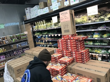 A person wearing a beanie and a dark jacket is stacking pizza boxes in the frozen food section of a grocery store. The shelves behind are filled with various packaged leafy greens, and there are signs indicating special prices. Several cardboard boxes and food products are visible around the person.