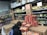 A person wearing a beanie and a dark jacket is stacking pizza boxes in the frozen food section of a grocery store. The shelves behind are filled with various packaged leafy greens, and there are signs indicating special prices. Several cardboard boxes and food products are visible around the person.