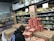 A person wearing a beanie and a dark jacket is stacking pizza boxes in the frozen food section of a grocery store. The shelves behind are filled with various packaged leafy greens, and there are signs indicating special prices. Several cardboard boxes and food products are visible around the person.