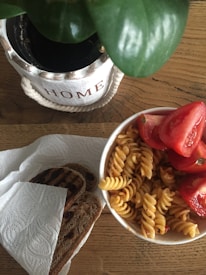 A wooden table holds a bowl of fusilli pasta mixed with sliced red tomatoes. Beside it, there is a piece of grilled bread placed on a paper towel. A potted plant in a white container with the word 'HOME' written on it is in the background.