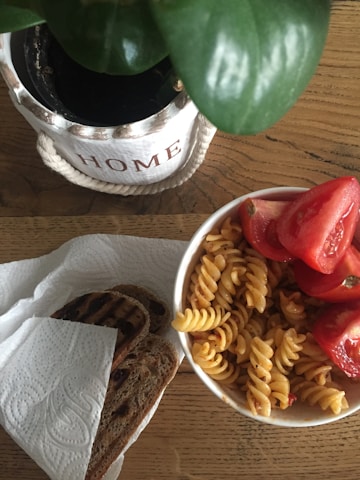A wooden table holds a bowl of fusilli pasta mixed with sliced red tomatoes. Beside it, there is a piece of grilled bread placed on a paper towel. A potted plant in a white container with the word 'HOME' written on it is in the background.