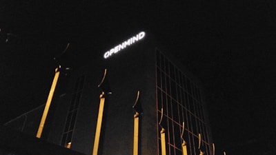 A building at night with a dark facade and a large white sign reading 'OPENMIND' illuminated on the upper section. The structure features several vertical golden-yellow elements resembling pillars or decorative fins against the dark background.