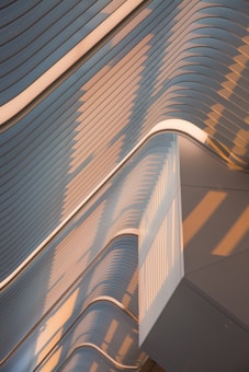 A modern architectural structure with flowing, curved lines and smooth surfaces. It features a pattern of horizontal slats with a blend of soft pastel colors, creating an impression of motion and fluidity. The lighting casts long shadows across the surfaces, enhancing the texture and depth of the design.