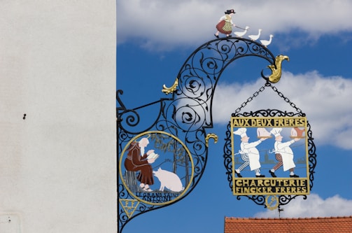 A decorative wrought iron sign hangs against a clear blue sky, featuring intricate designs with vibrant colors. One section of the sign depicts two chefs in white uniforms holding cooked meats, with the text 'AUX DEUX FRERES CHARCUTERIE FINCKER FRERES.' Another section shows a person in traditional clothing tending to geese, with a picturesque landscape in the background.
