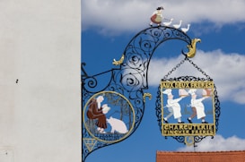 A decorative wrought iron sign hangs against a clear blue sky, featuring intricate designs with vibrant colors. One section of the sign depicts two chefs in white uniforms holding cooked meats, with the text 'AUX DEUX FRERES CHARCUTERIE FINCKER FRERES.' Another section shows a person in traditional clothing tending to geese, with a picturesque landscape in the background.