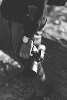 A person is holding a camera with both hands. The person is wearing a watch and has a tattoo on their arm. The image is in black and white, giving it a classic and timeless feel.