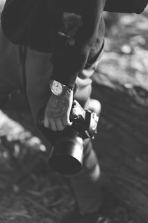 A person is holding a camera with both hands. The person is wearing a watch and has a tattoo on their arm. The image is in black and white, giving it a classic and timeless feel.