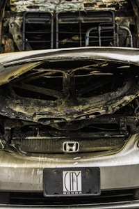 A heavily damaged car with noticeable burn marks, indicating it may have been involved in a fire or severe accident. The front grille is partially melted, and the hood is severely warped. A Honda badge is visible on the grille, and a license plate indicating the brand 'City Honda' is attached.