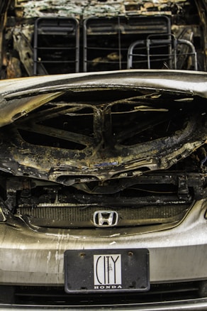 A heavily damaged car with noticeable burn marks, indicating it may have been involved in a fire or severe accident. The front grille is partially melted, and the hood is severely warped. A Honda badge is visible on the grille, and a license plate indicating the brand 'City Honda' is attached.