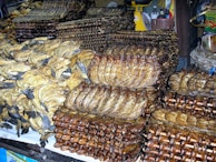 A market stall displays a large quantity of dried and smoked fish stacked in piles. The fish are arranged in various ways, some are flat and others are skewered, neatly organized into rows. The setting appears to be a busy market or shop with other goods visible in the background.