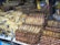 A market stall displays a large quantity of dried and smoked fish stacked in piles. The fish are arranged in various ways, some are flat and others are skewered, neatly organized into rows. The setting appears to be a busy market or shop with other goods visible in the background.