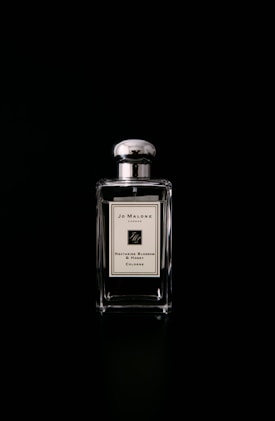 A clear glass bottle of Jo Malone Nectarine Blossom & Honey Cologne is centered against a black background. The bottle features a metallic cap and a minimalist label, giving it an elegant and luxurious appearance.