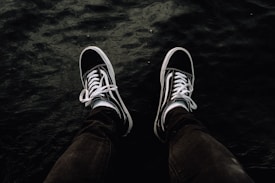 Two legs wearing dark sneakers dangle over a body of water, with the surface of the water appearing dark and somewhat textured.